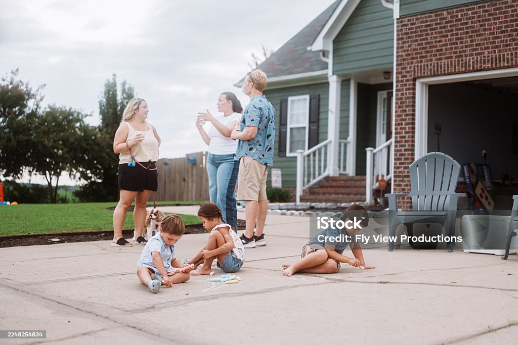 Kids sit on the driveway making chalk drawings while nearby adults talk casually during a relaxed neighborhood gathering.